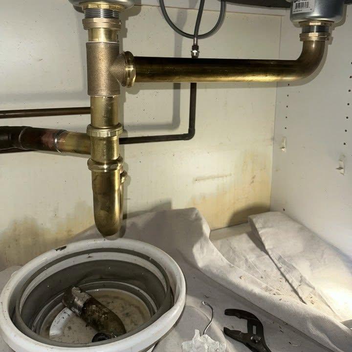 A brass double sink drain and trap under a kitchen sink.
