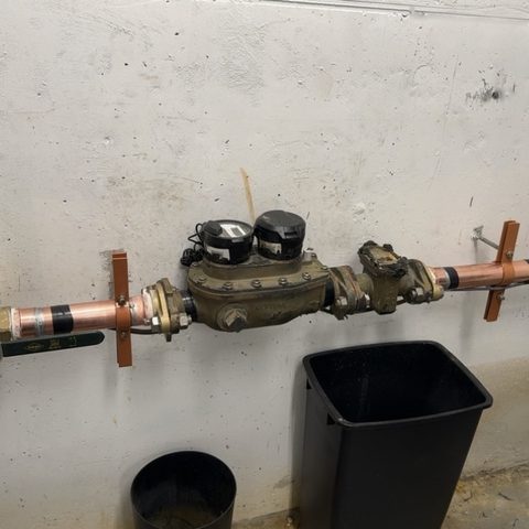A water meter installed on copper pipe along a concrete wall.