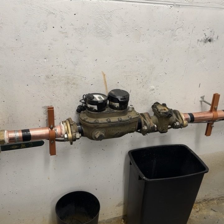 A water meter installed on copper pipe along a concrete wall.